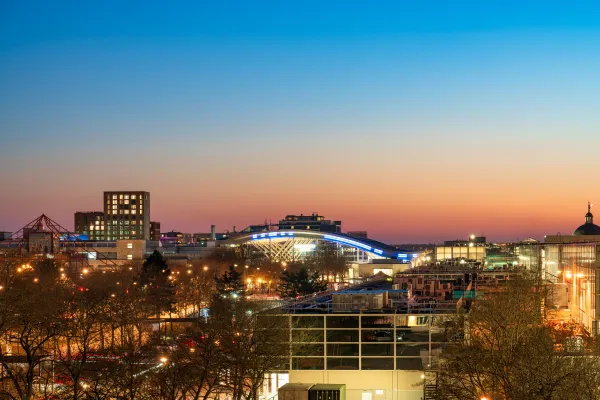 Urban cityscape at dusk with illuminated buildings and street lights, including a modern curved structure in the center, bare trees lining the streets, and a gradient sky transitioning from blue to orange above the roofline.