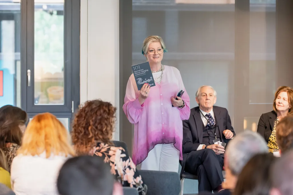 A woman in a pink cardigan speaks to an audience while holding a book, standing next to a man in a dark suit. Several audience members are visible in the foreground, seated and listening to the presentation in what appears to be a modern office or conference space.