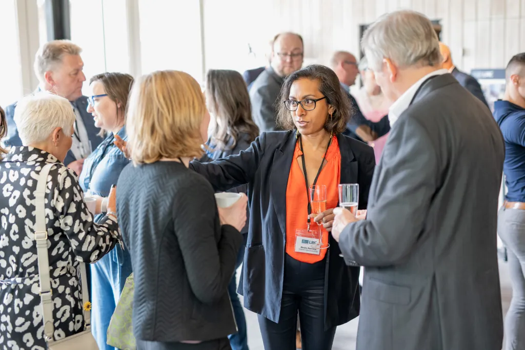 A woman wearing an orange conference badge and dark blazer engages in conversation with several attendees at a professional networking event, while other participants mingle in the background near large windows.