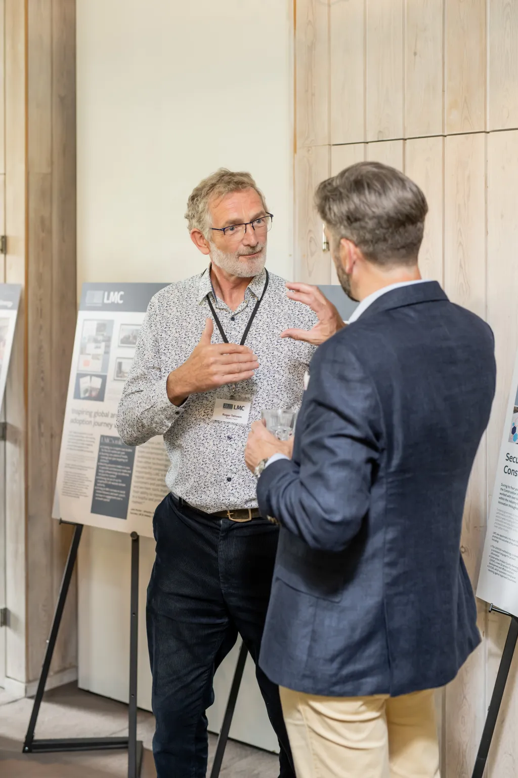 A man wearing glasses and a light patterned shirt discusses a poster presentation with another man in a dark blazer at what appears to be a professional conference or exhibition event.