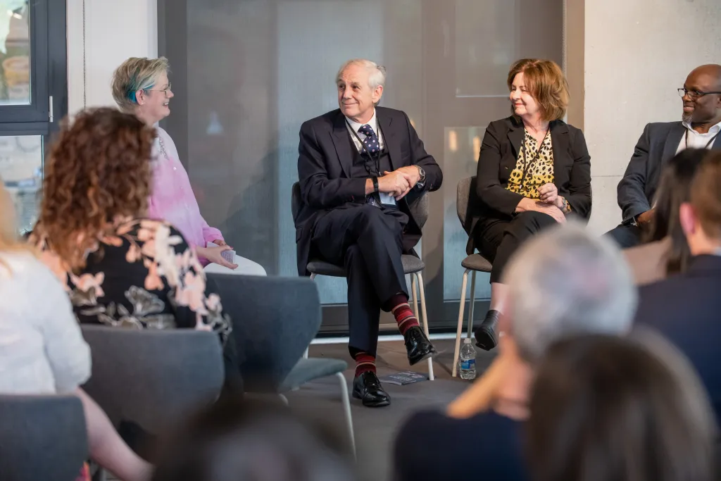Five professionals sit in a panel discussion format, with four panelists seated in the center facing an audience. The panelists include a man in a pink shirt, a man in a dark suit with a red tie, a woman in a yellow patterned top with a black blazer, and a man in a dark suit. Audience members are visible in the foreground, partially out of focus.