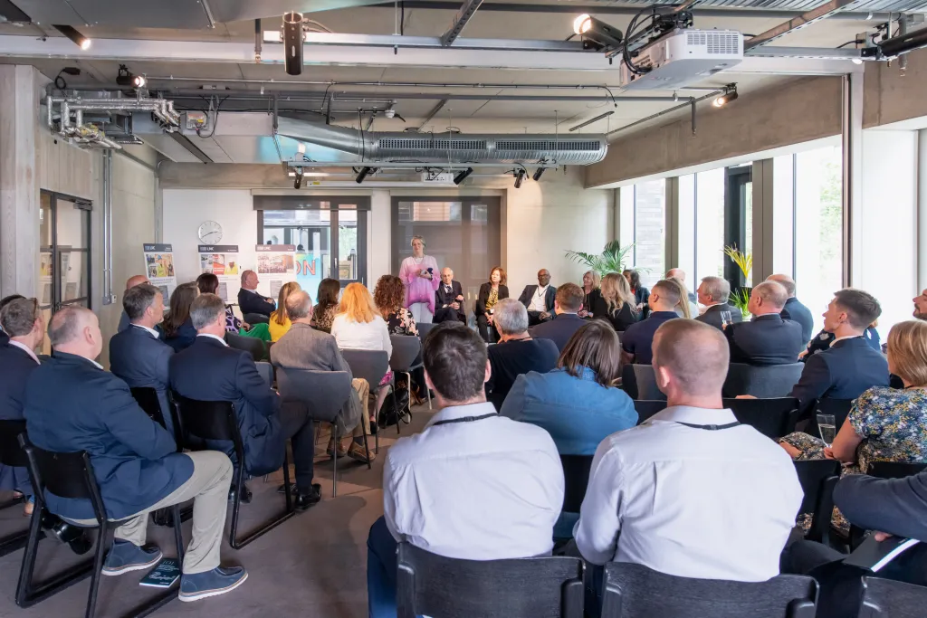 A diverse group of approximately 50 people gathered in a modern office space with floor-to-ceiling windows, seated and standing in a semi-circle facing toward the center where speakers are presenting. The attendees, dressed in business casual to formal attire, appear to be attending a company celebration or corporate event.