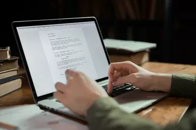 A person's hands are positioned over a laptop keyboard while reviewing a document displayed on the screen, with stacked books and a cup visible on the wooden desk beside the computer.