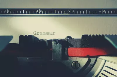 A typewriter with the word "Grammar" visible on the paper, with a ruler at the top and a red ribbon detail, symbolizing writing and language expertise.