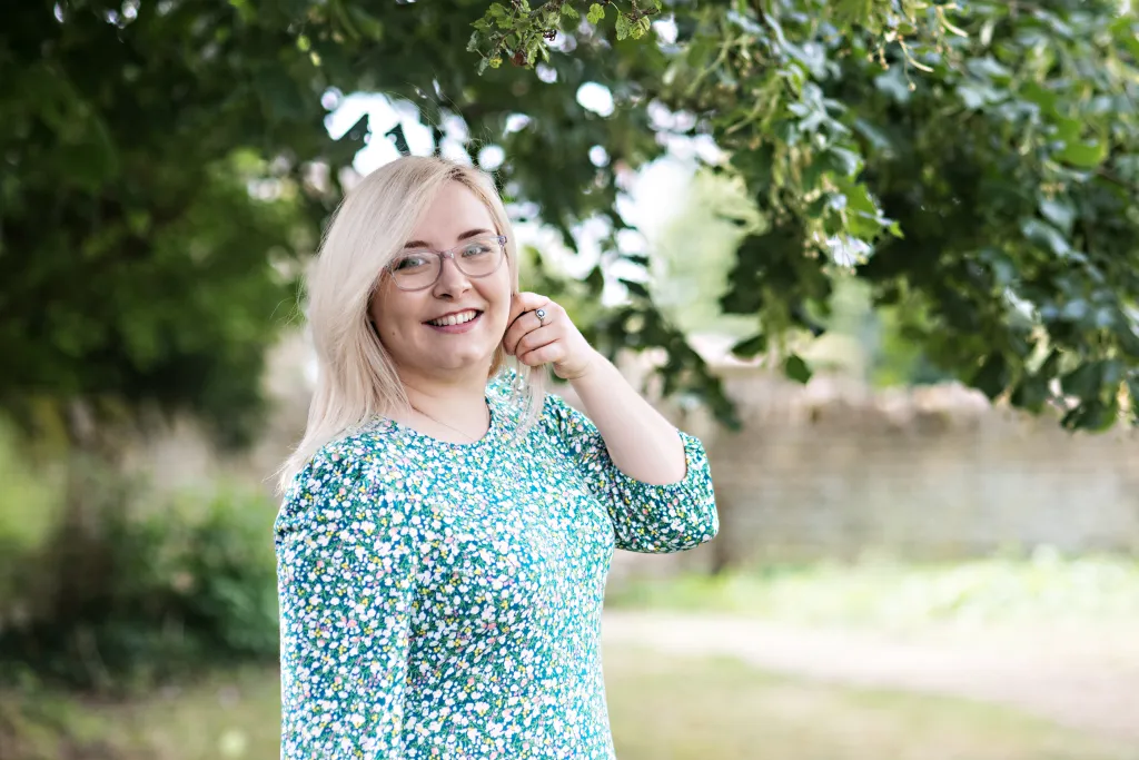 A woman with white hair and glasses wearing a turquoise floral top smiles at the camera while standing outdoors beneath tree branches.