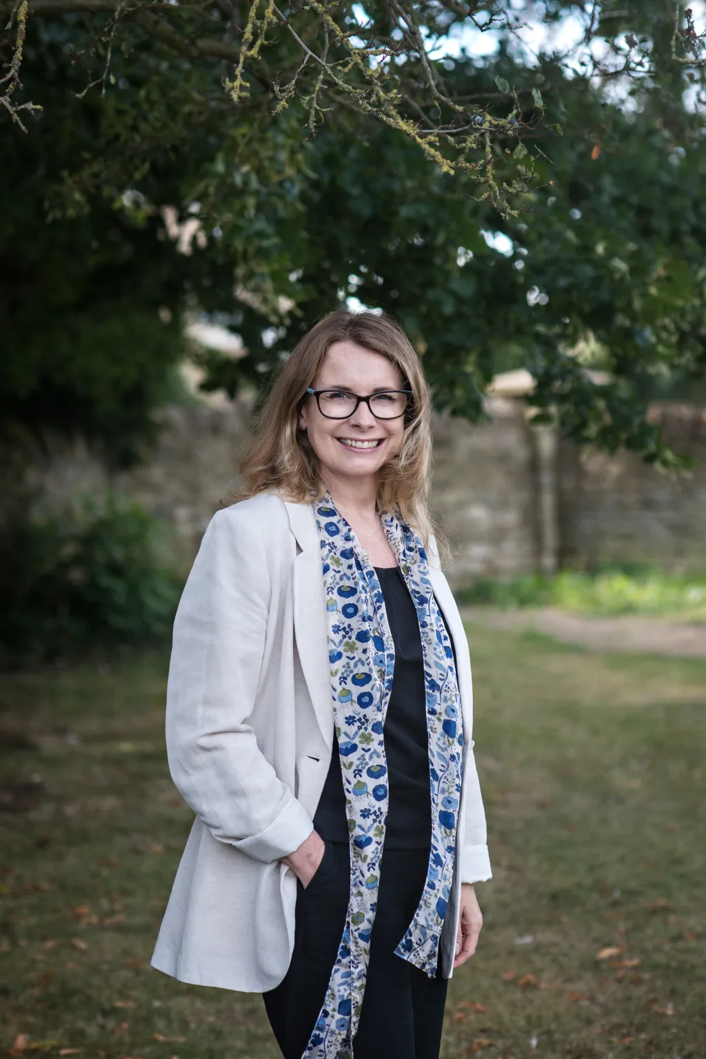 A woman with shoulder-length blonde hair wearing glasses stands outdoors under tree branches, dressed in a white blazer over a dark top with a blue patterned scarf. She smiles at the camera with her hand in her blazer pocket, with a stone wall visible in the blurred background.