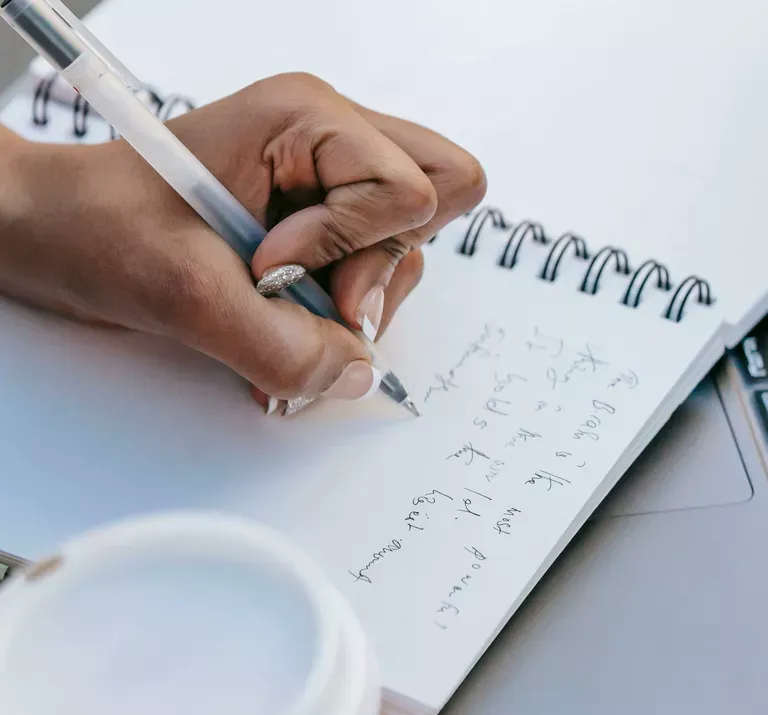 A person's hand holds a pen while writing mathematical equations in a spiral notebook on a white desk, with a coffee cup visible in the background.