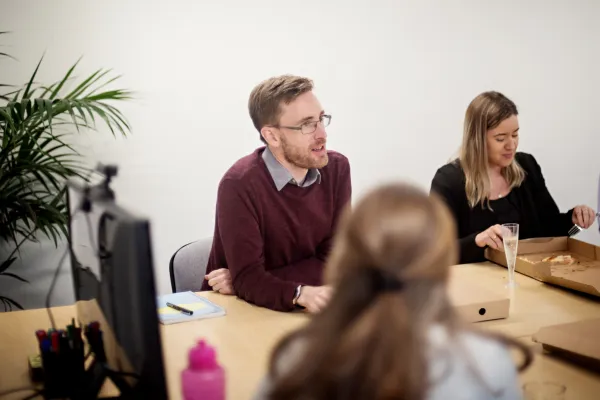 A man in a maroon sweater and glasses sits at a wooden table alongside a woman in a black top during what appears to be a professional meeting or interview. A person in the foreground is partially visible out of focus. The setting includes office elements like a potted plant, laptop, and desk accessories.