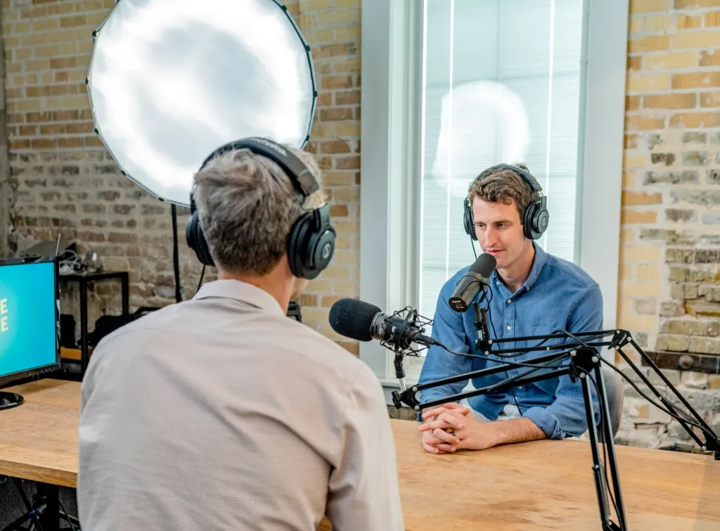 Two men conducting a podcast interview in a professional studio setting with brick walls, professional microphones, headphones, and a ring light.