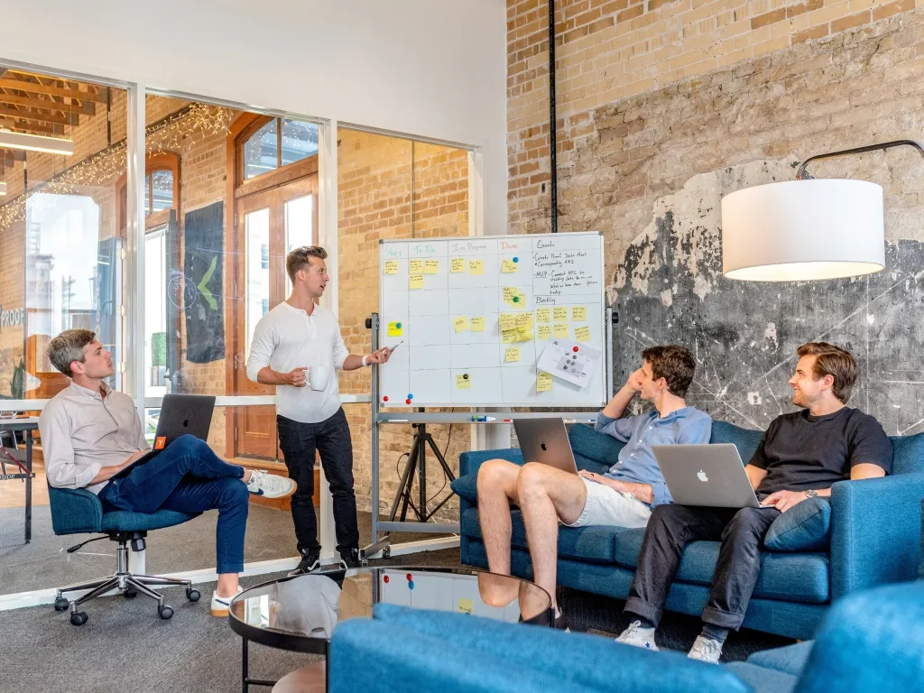 A diverse team of five people collaborates in a modern office space with exposed brick walls, gathered around a blue couch while one person stands presenting ideas on a whiteboard covered with yellow sticky notes. Laptops and casual seating create an informal brainstorming atmosphere with large windows and pendant lighting visible in the background.