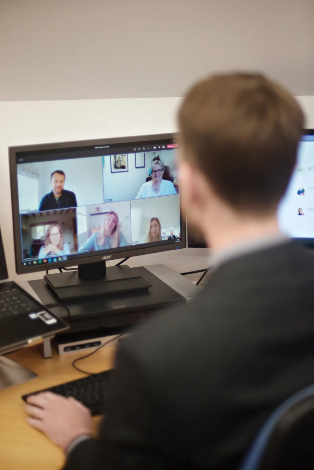 A person viewed from behind sits at a desk participating in a video conference call on a monitor, with multiple participants visible in a grid layout on the screen.
