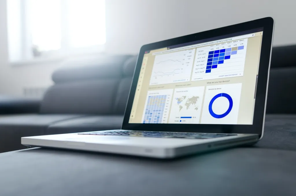 Laptop displaying a business analytics dashboard with multiple data visualization panels including charts, graphs, a world map, and a blue circular progress indicator on a desk.