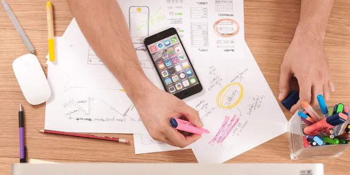 A designer's workspace from above with hands sketching wireframes and app layouts on papers scattered across a wooden desk, alongside a smartphone, mouse, and containers of colored markers and pens.