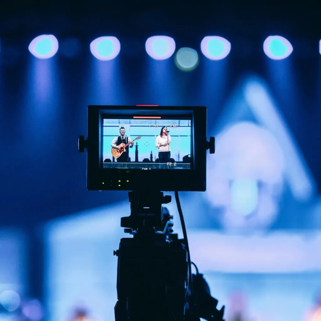 A camera monitor displays two performers on stage—one playing guitar and one singing—while blue spotlights illuminate the background during a live performance recording.