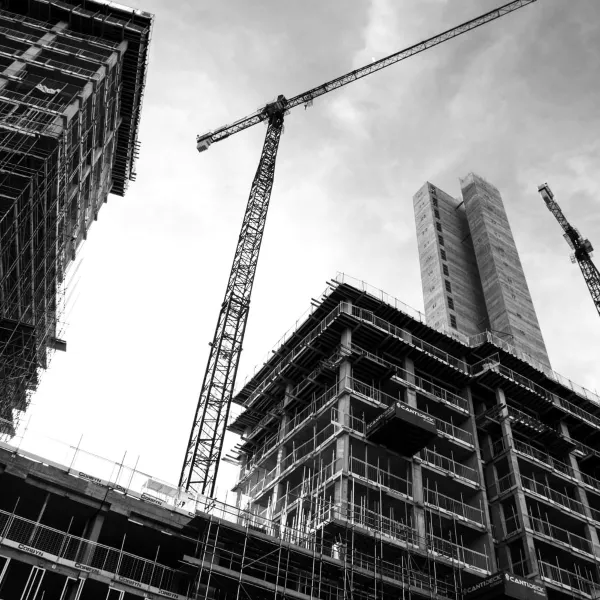 A construction site featuring multiple high-rise buildings under development, with a tall crane positioned centrally against a cloudy sky, concrete structures and scaffolding visible throughout the urban landscape.