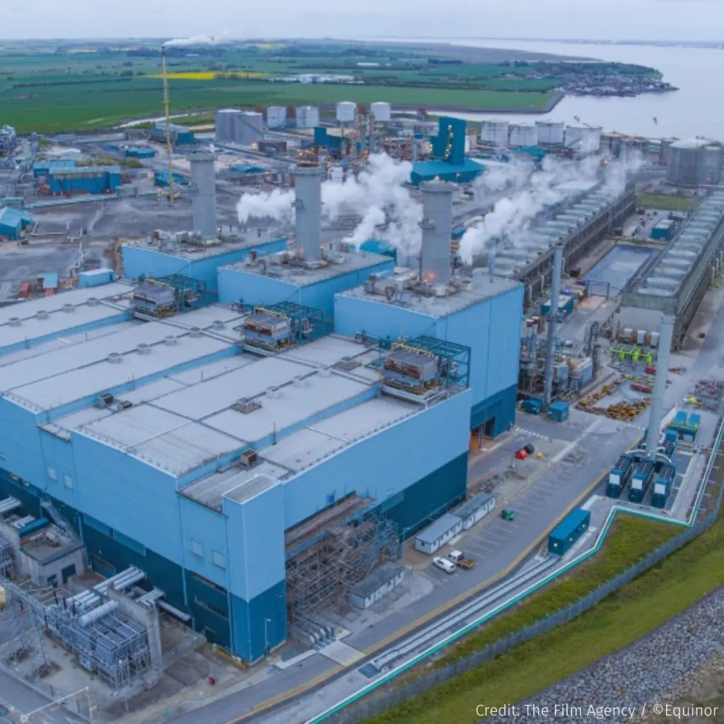 Aerial view of a large industrial manufacturing facility with blue-painted buildings, white roofed structures, and multiple smokestacks emitting steam, set in a coastal landscape with green fields and water visible in the background.