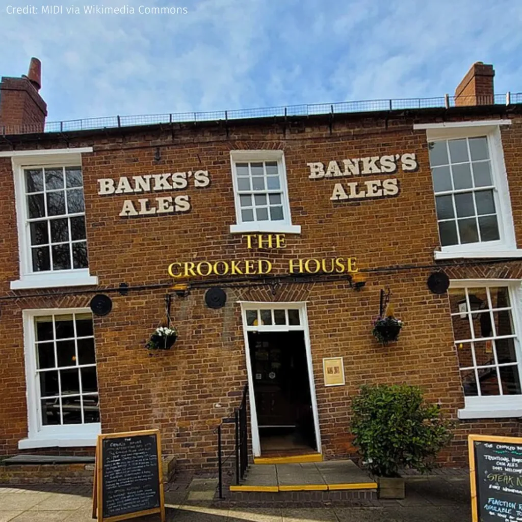 The Crooked House, a historic brick pub advertising Banks's Ales, displays noticeably tilted architecture with white-framed windows and a black door, exemplifying controversial building settling and structural issues.
