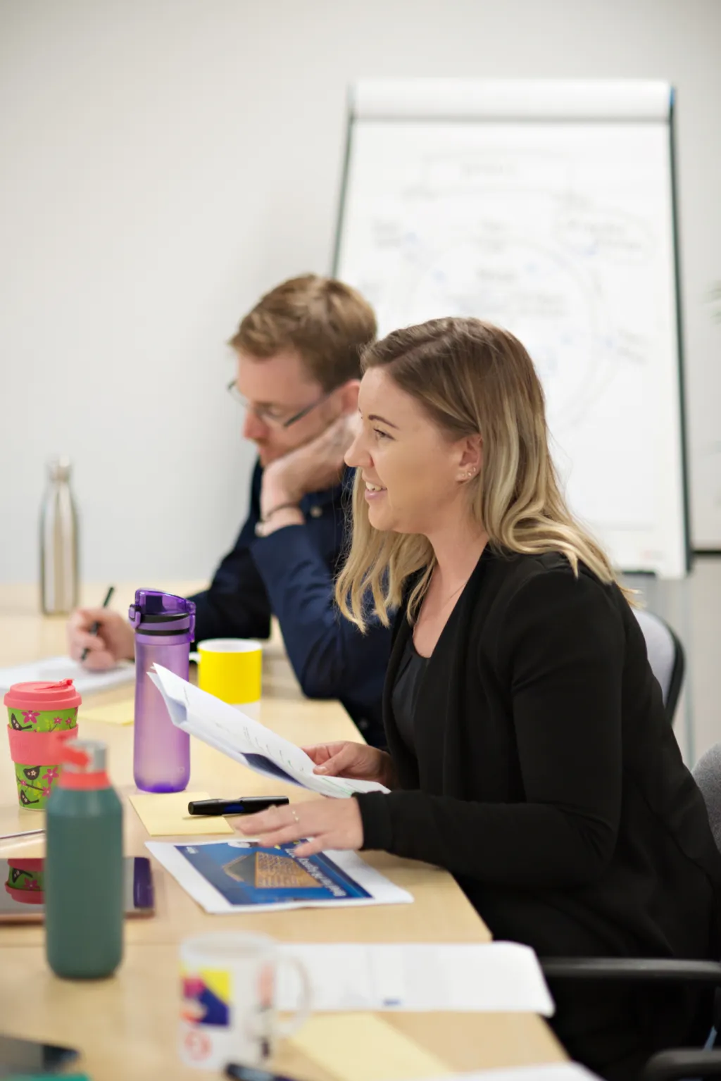 Two professionals sit at a table during a presentation or training session, with a whiteboard visible in the background. The woman in the foreground, wearing a black blazer, takes notes while smiling, and a man in a dark blue shirt sits beside her. Various materials and water bottles are arranged on the table in front of them.