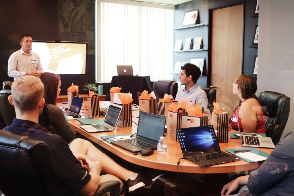 A man stands at the left presenting to a group of five people seated around a curved wooden table in a modern conference room. Laptops and popcorn containers are distributed across the table, with a presentation visible on a wall-mounted screen behind the presenter.