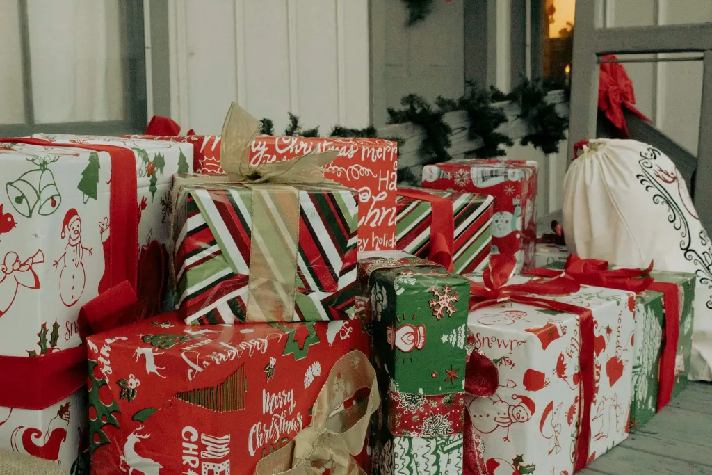 A collection of wrapped holiday gifts with red, green, and white festive paper stacked together on a porch, featuring Christmas-themed designs and patterns, accompanied by holiday garland decorations in the background.