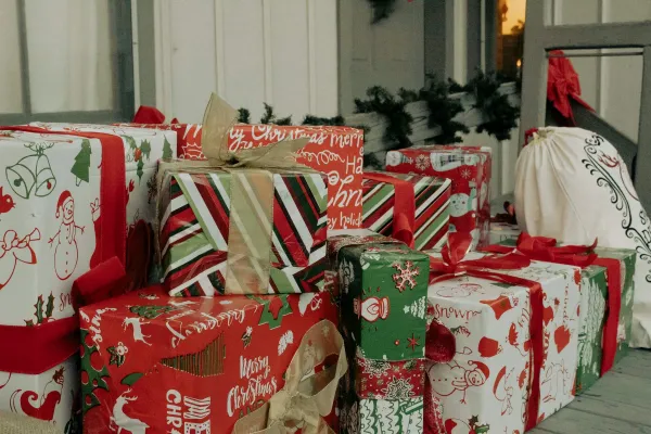 A collection of wrapped holiday gifts with red, green, and white festive paper stacked together on a porch, featuring Christmas-themed designs and patterns, accompanied by holiday garland decorations in the background.