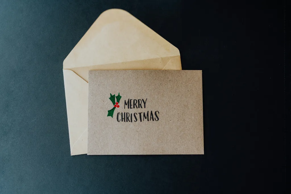 A kraft paper Christmas card with "MERRY CHRISTMAS" text and holly decoration lies on top of a cream-colored envelope against a dark teal background.