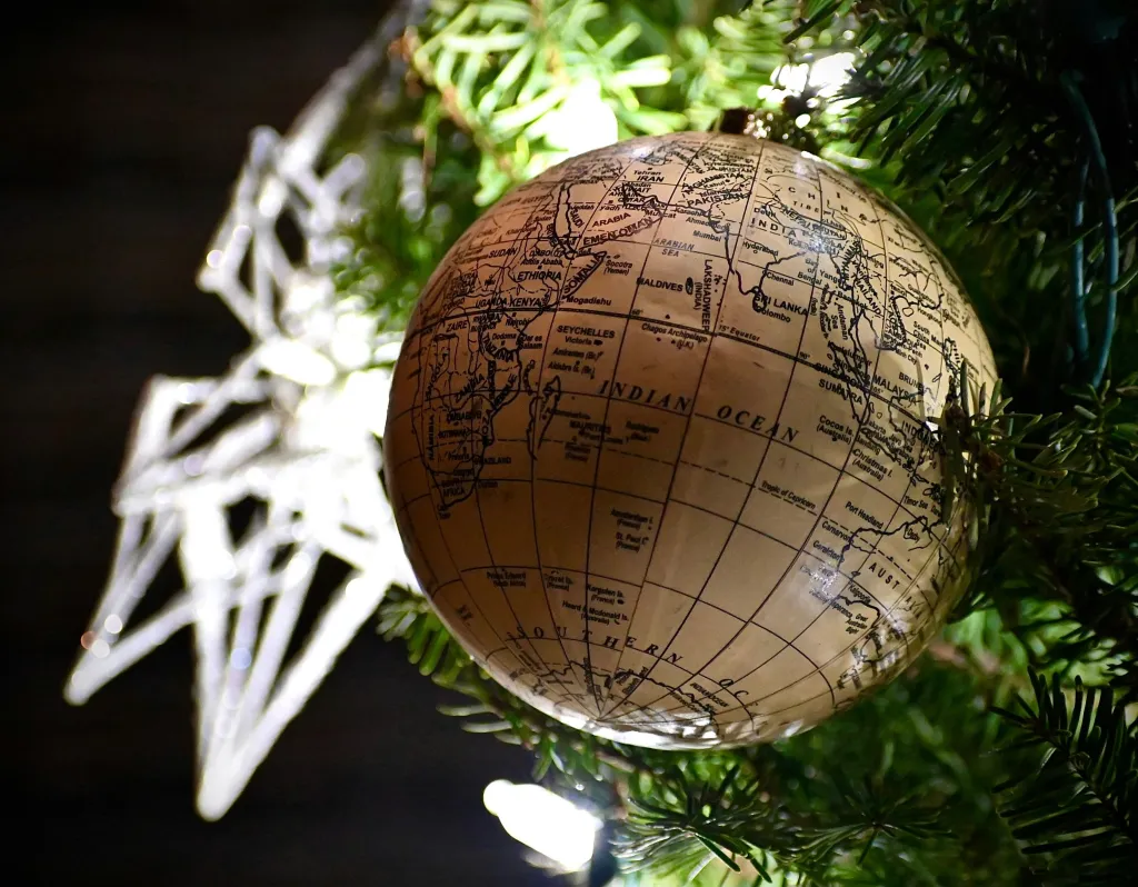 A wooden globe ornament with geographical markings hangs from an evergreen branch decorated with white star ornaments against a dark background, symbolizing global giving and holiday charity.