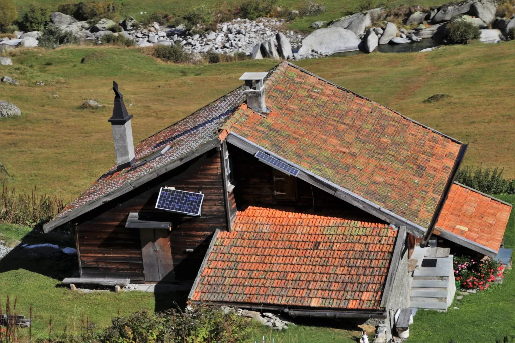 A rustic wooden chalet with terracotta tile roofs set on a grassy Alpine hillside, equipped with solar panels and a small external air conditioning unit, surrounded by rocky terrain and vegetation.