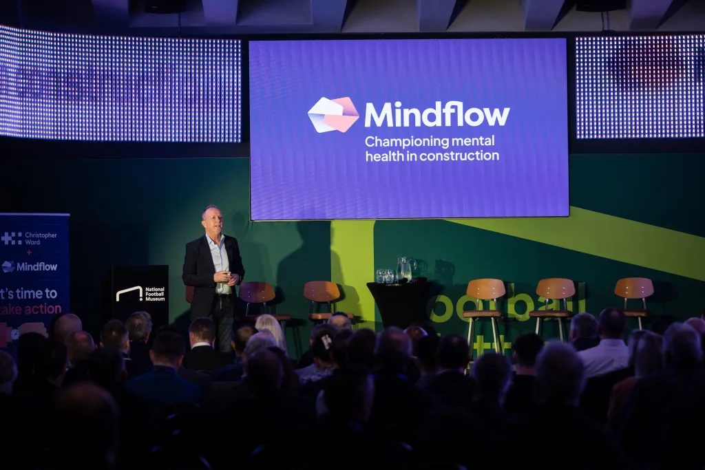 A speaker stands on stage presenting to an audience with a large purple screen behind him displaying the Mindflow logo and tagline 