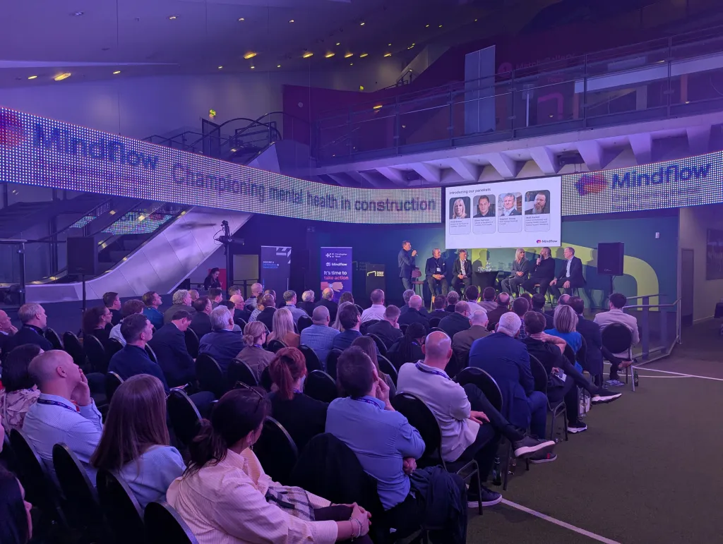 A large indoor event with an audience seated in rows facing a stage, illuminated by purple and blue lighting. Several speakers stand on the stage addressing the crowd, with branded banners and screens visible in the background. The venue appears to be a professional conference or seminar space focused on a mental health initiative for construction workers.