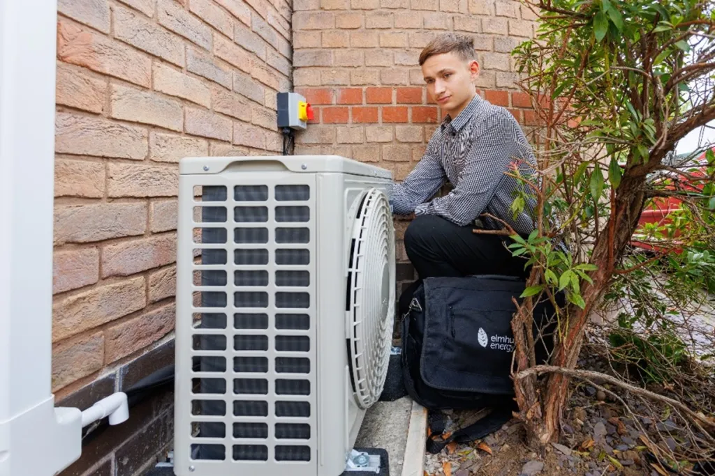 A technician in a dark jacket crouches beside an outdoor air heat pump unit mounted on a brick wall, inspecting or servicing the equipment as part of green building installation and maintenance work.