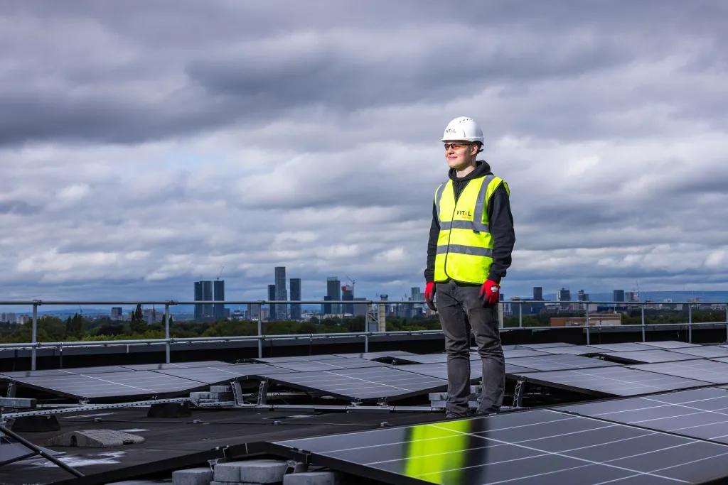 A construction worker wearing a white hard hat and high-visibility yellow vest stands on a rooftop among solar panels, with a city skyline visible in the background under a cloudy sky.
