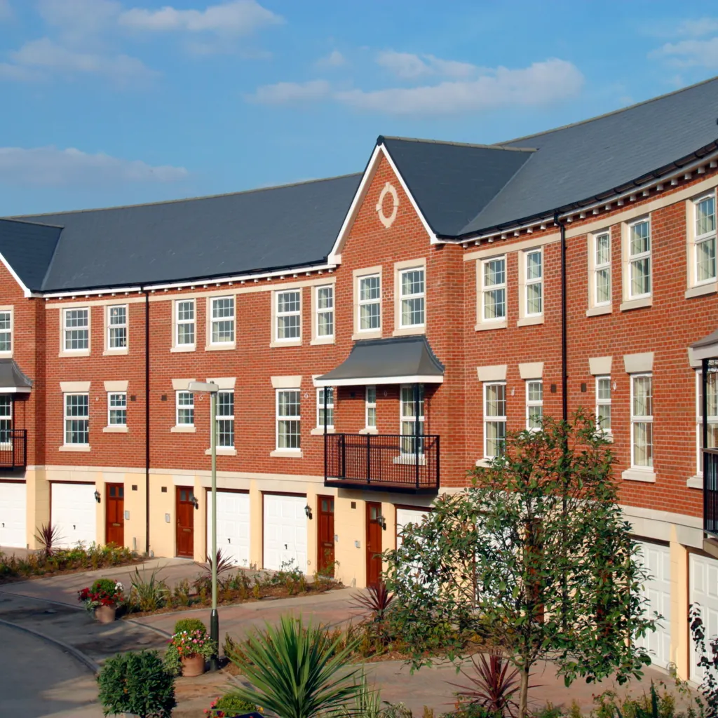 A modern brick residential building with symmetrical architecture, featuring multiple windows with white frames, a central peaked gable, and ground-level garages with red doors fronting a paved courtyard with landscaping.