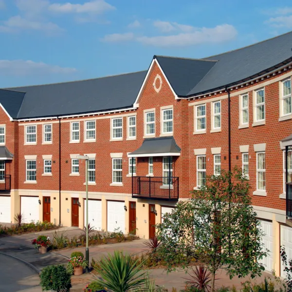 A modern brick residential building with symmetrical architecture, featuring multiple windows with white frames, a central peaked gable, and ground-level garages with red doors fronting a paved courtyard with landscaping.