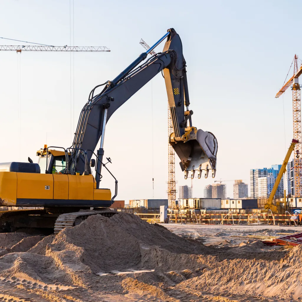 A large yellow and dark blue excavator operates on a sandy construction site, with its bucket raised high. Multiple construction cranes and a city skyline are visible in the background, indicating an active urban development project.
