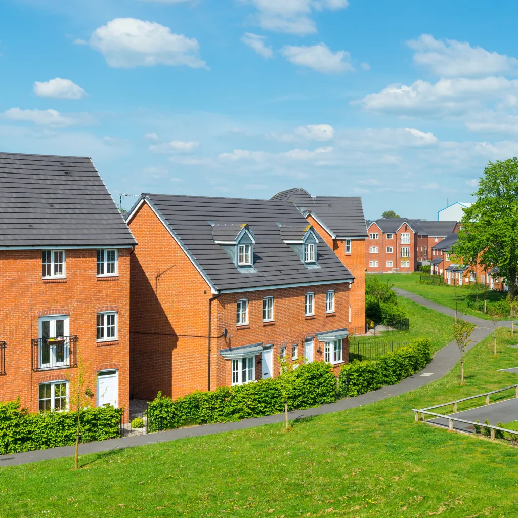 A modern residential development of brick-built houses with dark slate roofs arranged along a curved street, set on a sloping green lawn under a blue sky with white clouds.