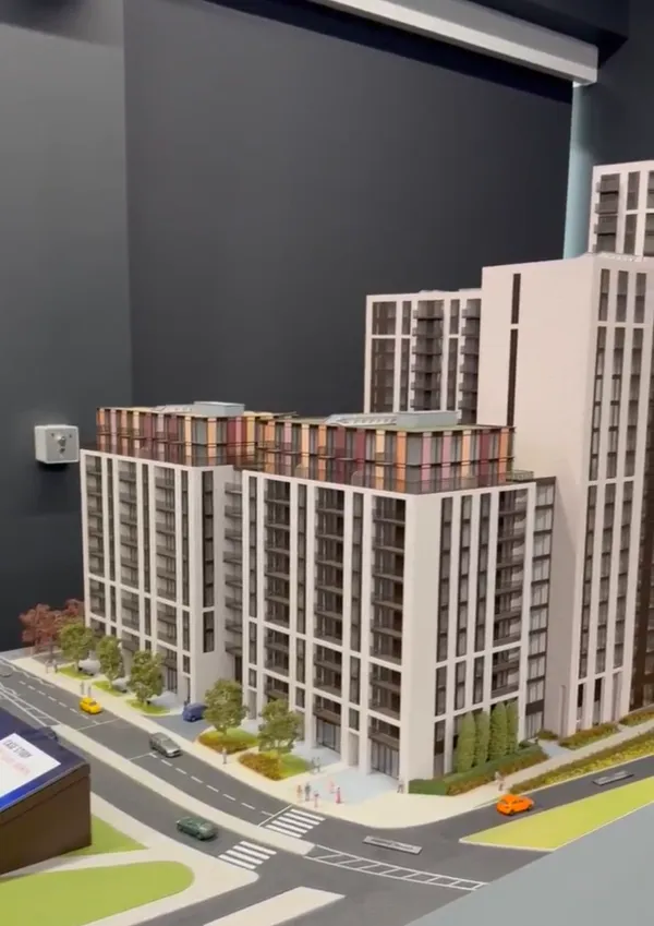 Architectural model of a contemporary mixed-use development featuring multiple residential buildings with brick and white facades arranged around a street-level plaza with green spaces and pedestrian walkways.