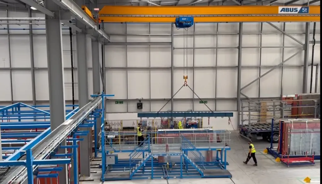 A large industrial warehouse with an overhead crane lifting a prefabricated building component, while a worker in a yellow safety vest supervises from the floor below alongside stacked construction materials and blue structural frameworks.