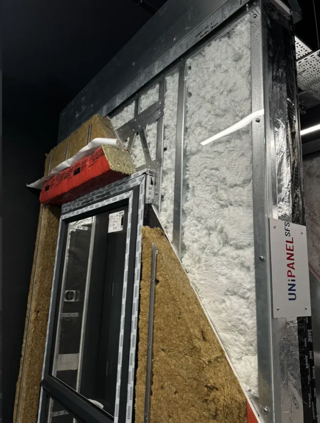 A prefabricated building module under construction showing insulation layers, including mineral wool and foam boards, with an installed window unit and red beam visible at the top, demonstrating modern offsite construction methods.