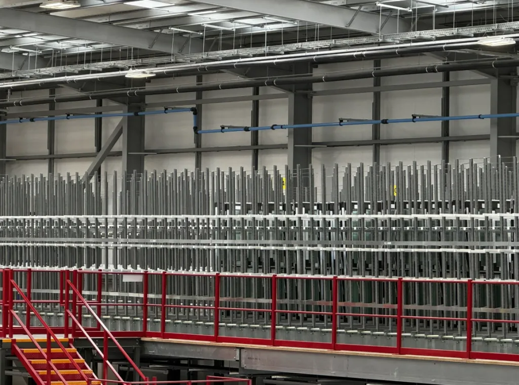 A large industrial warehouse with metal framework and white vertical posts arranged in rows, featuring red safety railings and stairs in the foreground, and fluorescent ceiling lights illuminating the spacious construction facility.