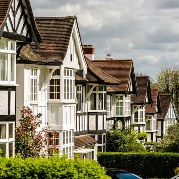 A row of Tudor-style houses with distinctive black and white timber framing, steeply pitched roofs, and large windows line a tree-lined street. The homes feature traditional architectural details including decorative half-timbering and terracotta roof tiles. Manicured hedges and vegetation frame the residential properties under a partly cloudy sky.