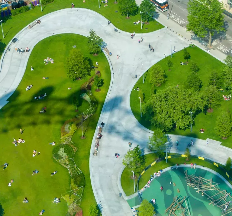 Aerial view of a modern urban park with curved pathways, green lawns, scattered trees, and people relaxing in small groups. A turquoise water feature or pool is visible in the lower right corner. The design shows accessible, interconnected spaces that encourage community gathering.