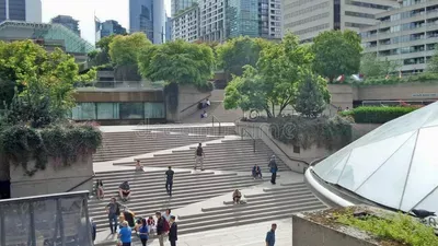 A wide outdoor plaza with multiple levels of seating steps where diverse groups of people gather and socialize. Modern buildings and green trees surround the space, with a distinctive white curved architectural element visible on the right side.