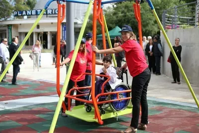 Two adults in red shirts assist a child in a wheelchair using an accessible playground swing structure with yellow and red frame elements, while other visitors explore the inclusive playground in the background.