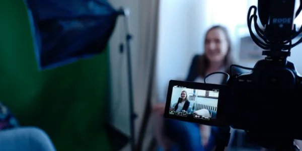 A woman sits in front of a professional camera setup with a green screen backdrop while being recorded for a live stream broadcast.