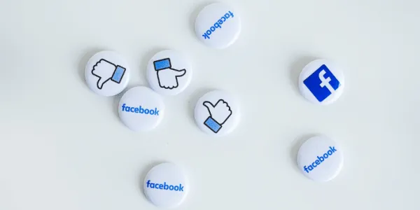 Several white and blue circular tokens scattered on a light background, each displaying the Facebook logo, thumbs-up icon, or the word 