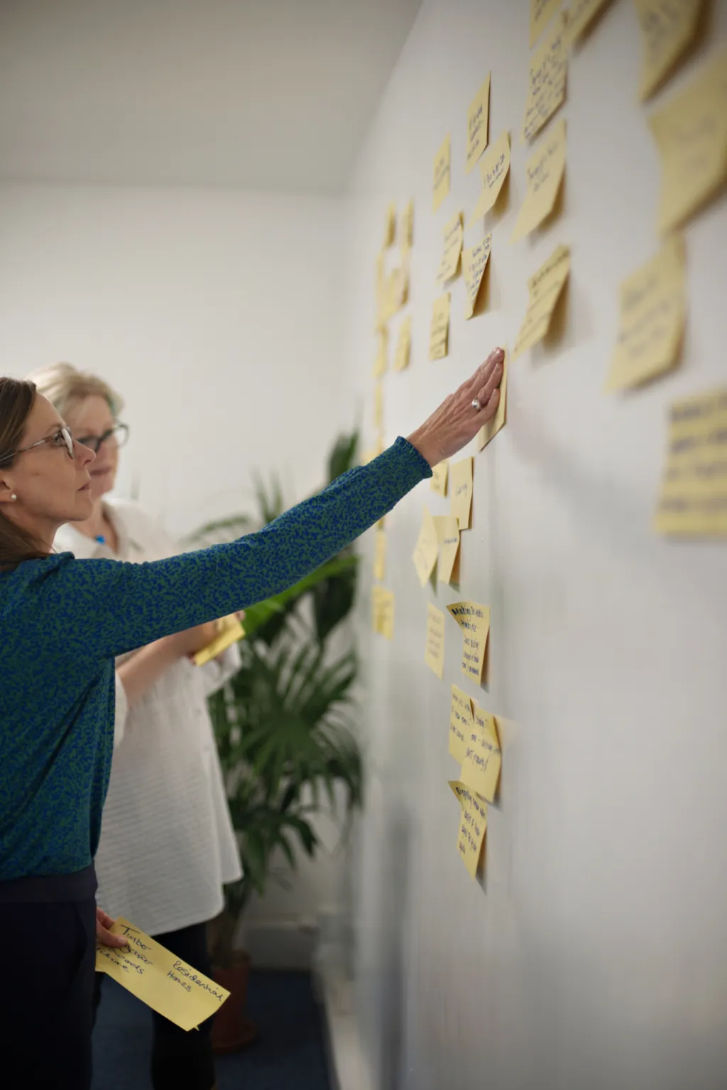 A woman wearing glasses and a teal cardigan reaches up to point at sticky notes arranged on a white wall, with a plant visible in the background, during what appears to be a brainstorming or planning session.