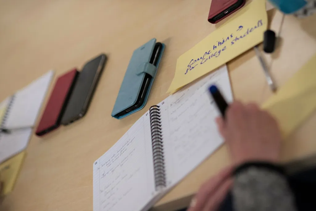 A person's hand writes notes in a spiral notebook surrounded by colorful pens and sticky notes on a beige surface, illustrating planning or brainstorming for content creation.