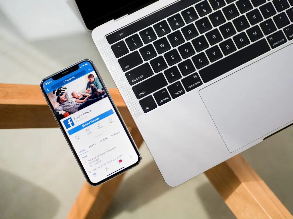 A smartphone displaying a Facebook post with a video thumbnail sits next to a laptop on a wooden desk, illustrating live streaming setup for content creators.