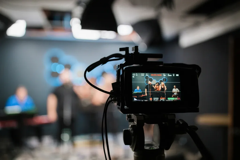 A camera mounted on a tripod displays a live feed on its screen showing a studio setup with people and blue lighting in the background, capturing content for live streaming.